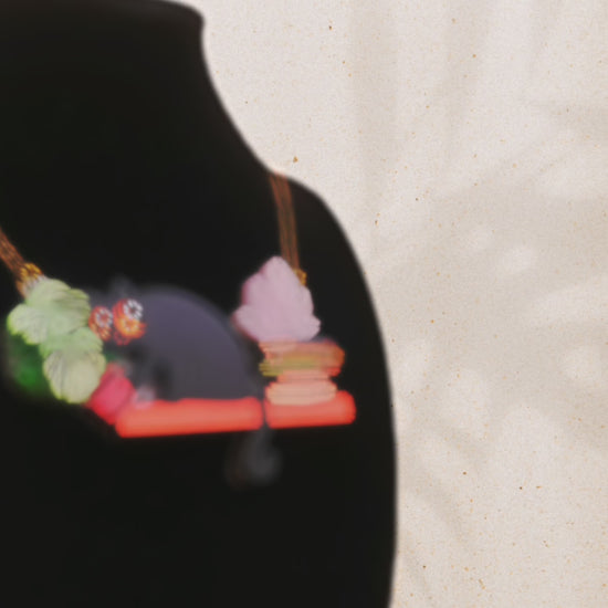 Video of front and back of mixed material handmade necklace of chibi cartoon cat sat on a witch's book shelf with pearlescent crystal. knocking a plant pot of the edge, with a gold chain and black gift box with a blue familiars collection gift sleeve. on black bust.